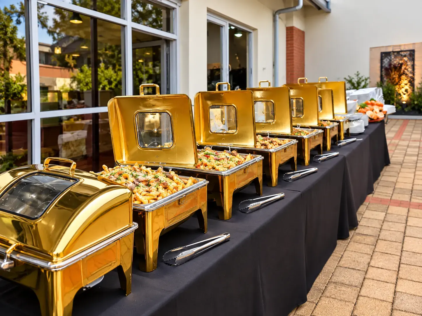 A long buffet line of gold chafing dishes on a courtyard terrace, lids open over pasta and savoury dishes, with serving tongs laid ready
