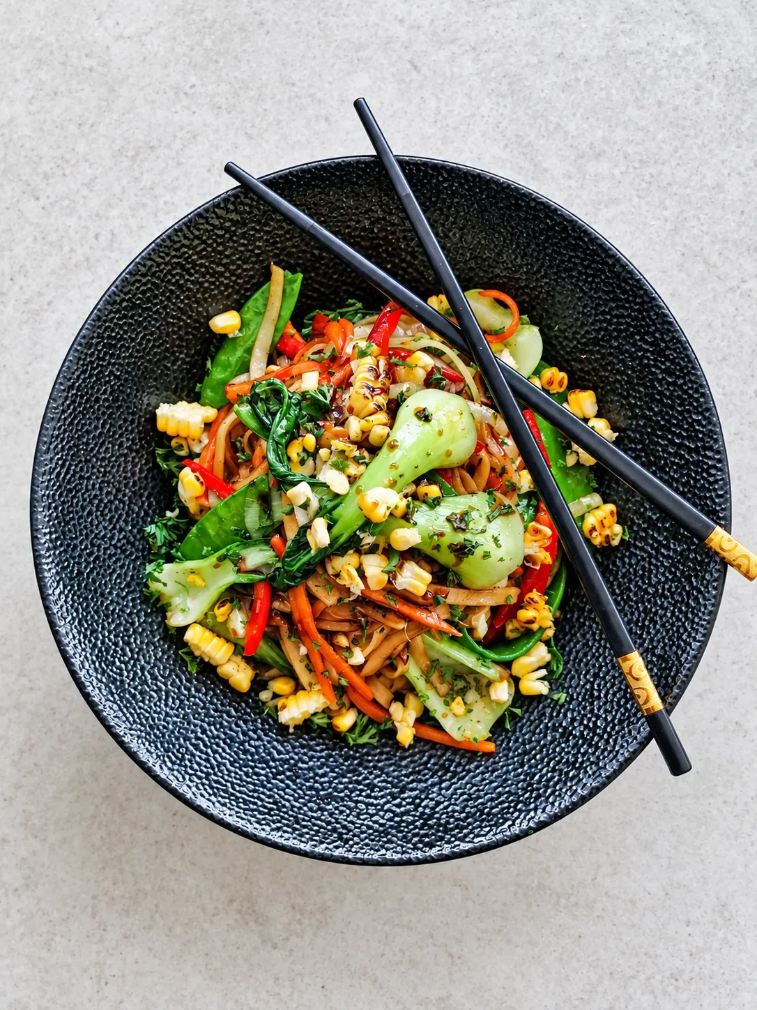 A vegetable stir-fry of noodles, bok choy, sugar snap peas, peppers and charred corn in a black bowl with chopsticks