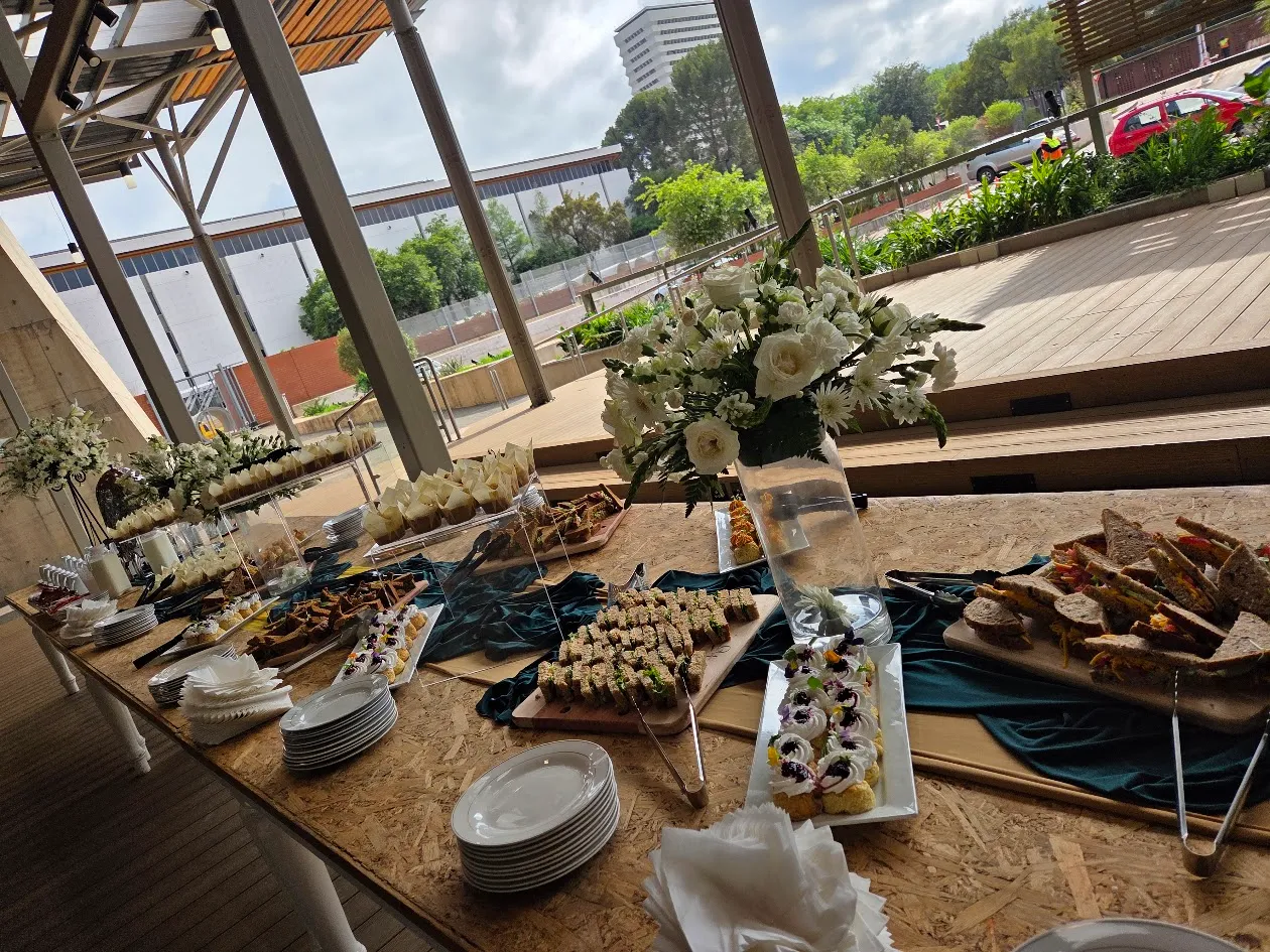 Elegant catering table setup with assorted gourmet finger foods, desserts, and sandwiches, styled with white floral arrangements at a modern outdoor event venue.